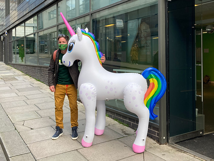 On the pavement in front of The Ledward Centre, a gentleman wearing a protective mask stands next to a large inflatable unicorn with a pink horn and a rainbow-coloured mane and tail.