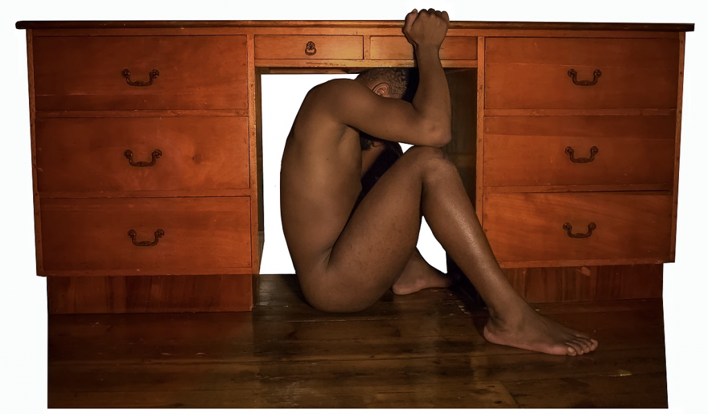 A naked young man with brown skin crouches side-on under a teak desk.