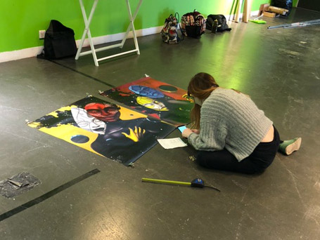 A woman sits on the floor surrounded by a measuring tape and two paintings.