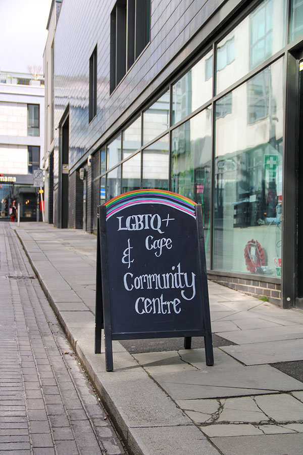An A-Board advertising on Jubilee Street outside the Ledward Centre entrance. LGBTQ+ Cafe & Community Centre is written in white on black. The arched top of the board is decorated with a chalk rainbow.
